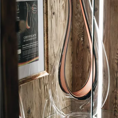 Eine elegante Glasvase mit einem modernen Design steht in einer Vitrine. Neben der Vase sind ein Zertifikat und einige Weingläser zu sehen.