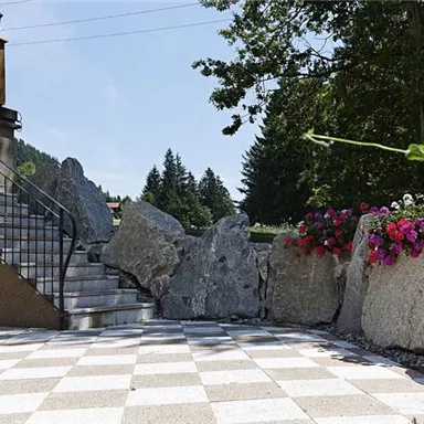 A paved outdoor area with a staircase and large stones. Colorful flowers grow along the rocks and there are trees in the background.