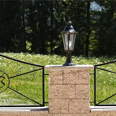 An elegant lantern stands on a stone pillar next to a wrought-iron fence. In the background, a green meadow with flowers can be seen.