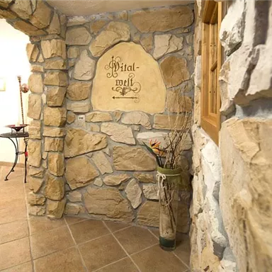 A rustic entrance area with stone walls and a decorative inscription. The floor is tiled and there are some plants and furniture pieces in the room.