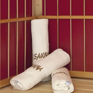 Three clean, white sauna towels are lying on a wooden bench in a sauna room. The walls are painted red, creating a relaxing atmosphere.