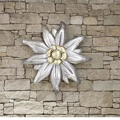 An artistic wall decoration in the form of a flower made of silver is placed in front of a stone wall. The flower has a central golden blossom and radiates elegance.