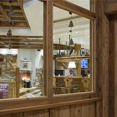 A cozy restaurant with wooden decor and warm lighting. The windows offer a view of cozy seating areas and a bar.
