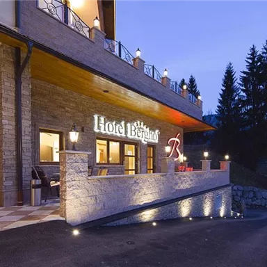 A modern hotel building with an inviting entrance area. The facade is made of stone and wood, surrounded by trees and gentle lighting.