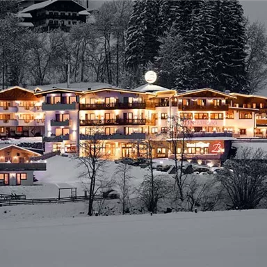 A illuminated hotel in the wintry snow. The surroundings are quiet and idyllic with trees in the background.