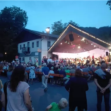 A lively outdoor event with a large crowd watching a concert on a stage. The evening sky is blue and the stage is beautifully lit.