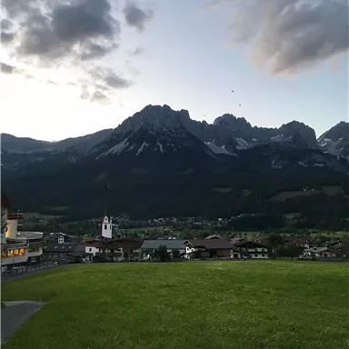 Eine beeindruckende Berglandschaft bei Sonnenuntergang mit einem kleinen Dorf im Vordergrund. Die Wolken sind sanft und die Berge sind teilweise schneebedeckt.