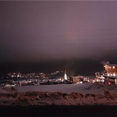 Eine schneebedeckte Landschaft bei Nacht mit Lichtern in der Ferne. Im Vordergrund stehen Gebäude und eine Kirche, umgeben von Schnee.