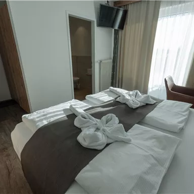 Ein modernes Hotelzimmer mit einem großen Bett und weißen Kissen. Im Hintergrund gibt es einen Fernseher und viel Tageslicht durch die Fenster.