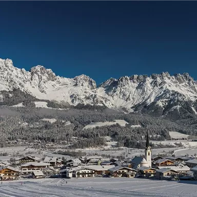 A snowy mountain landscape with high peaks and a picturesque village in the foreground. The sky is clear and blue.
