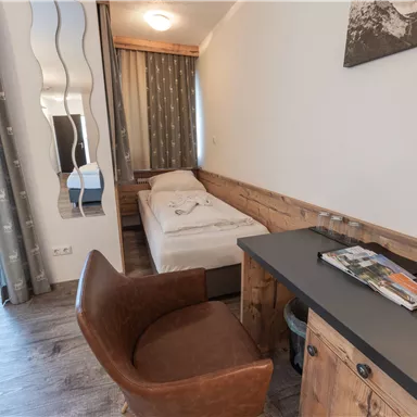 Ein helles Zimmer mit einem Einzelbett und modernen Möbeln. An der Wand befinden sich Holzverkleidungen und ein kleiner Schreibtisch mit einem Stuhl.