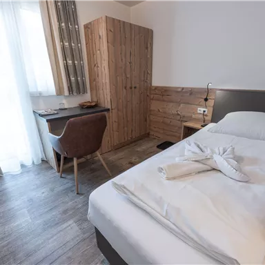 Ein modernes Einzelzimmer mit Holzmöbeln und einem gemütlichen Schreibtisch. Tageslicht strömt durch große Fenster und sorgt für eine einladende Atmosphäre.