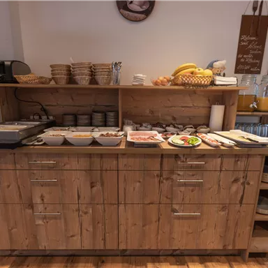 Ein Frühstücksbuffet mit einer Holzküste, die verschiedene Speisen wie Aufschnitt, Käse und Obst präsentiert. Im Hintergrund sind Teller und Schüsseln ordentlich angeordnet.