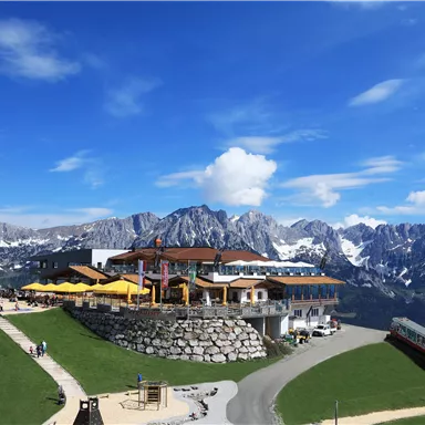Eine malerische Berghütte mit Terrasse und Sonnenschirmen in den Alpen. Im Hintergrund erheben sich majestätische Berge unter einem klaren blauen Himmel.