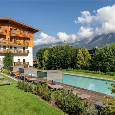 A beautiful hotel with a pool and loungers in the garden. In the background, there are mountains and a blue sky.