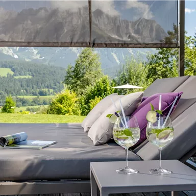 A cozy lounger with cushions and two cocktails on a table. In the background, green landscape and mountains can be seen.