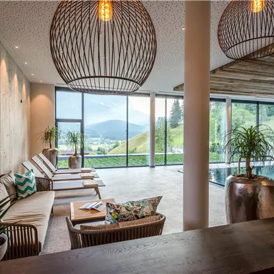 A modern wellness area with comfortable loungers and large windows. In the background, a green landscape can be seen.