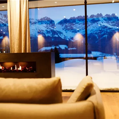 A cozy living room with a modern fireplace. Through large windows, one can see snow-covered mountains and a clear evening sky.