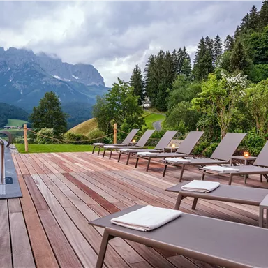 An elegant outdoor area with sun loungers by the pool. In the background, majestic mountains and green forests can be seen.