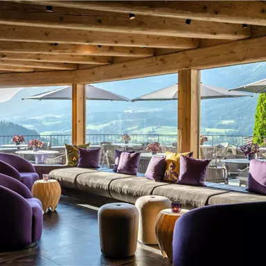 An elegant lounge area with modern furniture and large windows. The view of the mountains and the sun creates a cozy atmosphere.