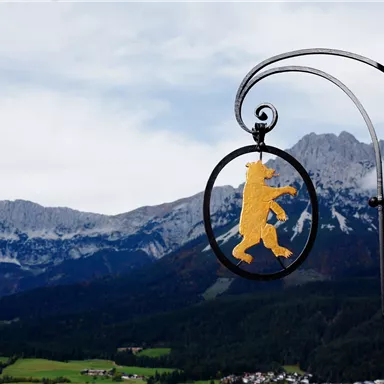 A golden bear hangs from an iron ring in front of an impressive mountain landscape. The mountains are covered with clouds, and the nature is green.