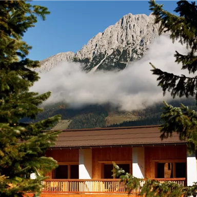 A beautiful mountain panorama with a majestic peak and clouds. In the foreground, there is a wooden building with balconies and trees.