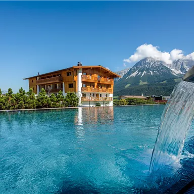 A luxurious pool with clear water and a waterfall. In the background, mountains and a traditional wooden house can be seen.