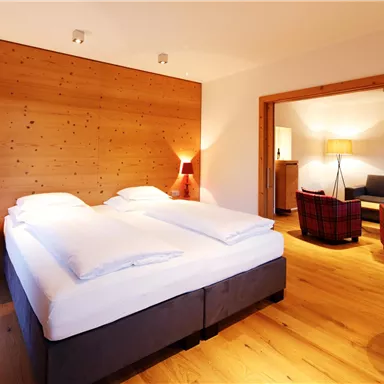 A modern bedroom with a large double bed and wooden walls. In the background, there is a cozy seating area with a sofa and two armchairs.