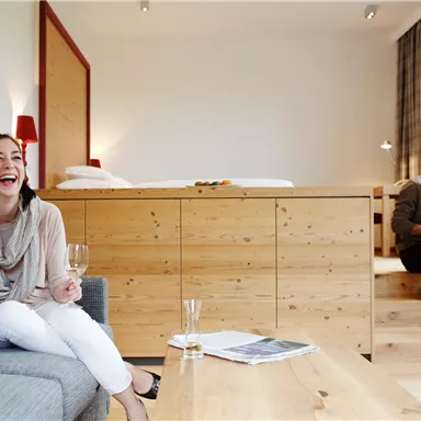 A woman is sitting and laughing on a sofa, holding a glass in her hand. In the background, a man can be seen sitting at a table.