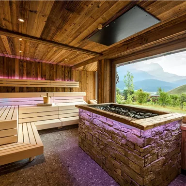 A cozy sauna room made of wood with a stone wall and colorful lights. Large windows provide a view of the mountains and nature.