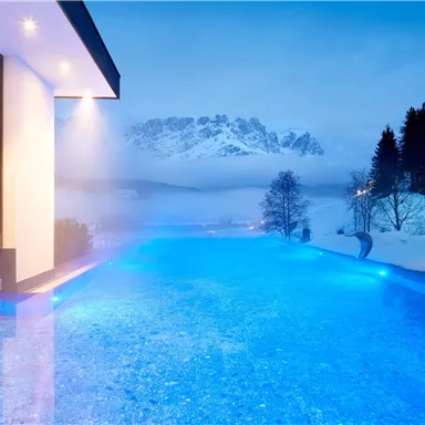 A modern house with a blue infinity pool and snow-covered mountains in the background. The landscape is calm and wintry, with a blurred view of the mountains.
