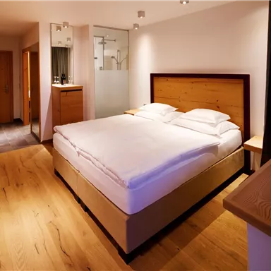 A modern bedroom with a large bed and wooden floor. The walls are bright and there is a small area with a shower in the background.