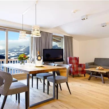 A modern living room with a dining table, chairs, and a television. Large windows offer a view of the mountain landscape.