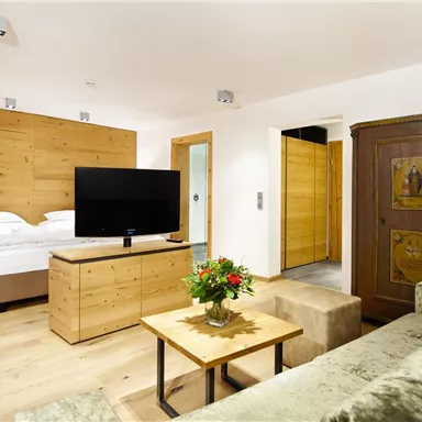 A modern hotel room with wooden decor and a cozy sofa. The space includes a bed, a television, and a small table with flowers.