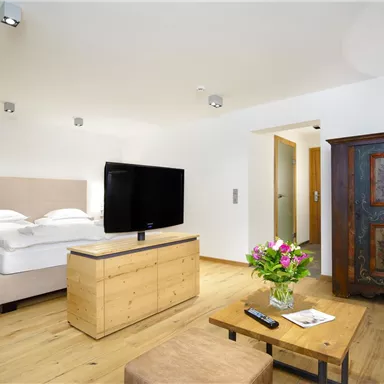 A modern bedroom with a double bed, a television, and a cozy seating area. The room is bright and features wooden flooring as well as a beautiful flower decoration.