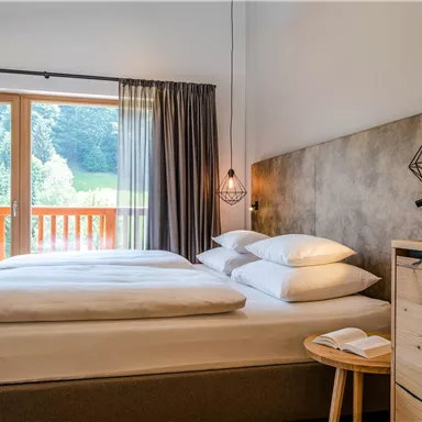 A modern bedroom with a large double bed and wooden decor. The windows offer a view of the green mountains.