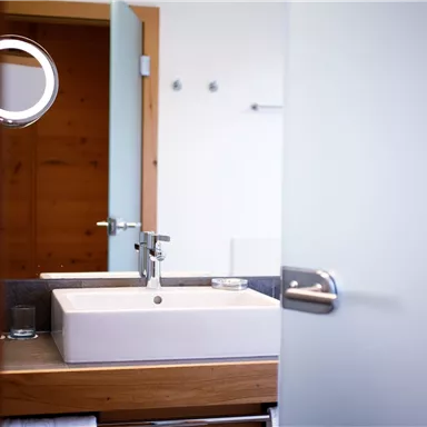 A modern bathroom with a white sink and a wooden shelf. The door is open and lets light in.