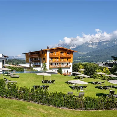 A picturesque hotel in the mountains with a large garden. The lawn is equipped with deck chairs and sun umbrellas, while the mountains are visible in the background.