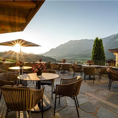 A cozy terrace with tables and chairs, surrounded by mountains. The sun shines bright and creates an inviting atmosphere.