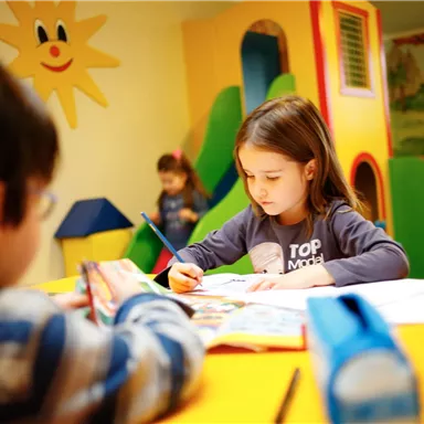 Two children are playing in a colorful room. A girl is painting intently at the table, while a boy is busy in the background.