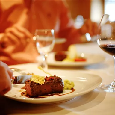A set table with a dish of beef and side dishes. In the background, there are wine glasses and people enjoying their meal.