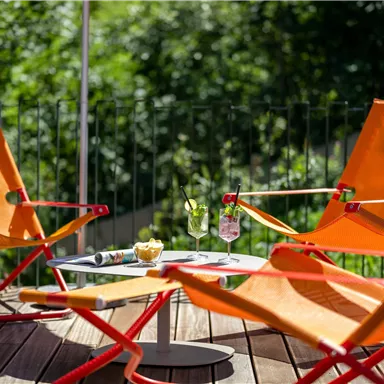 Two orange lounge chairs are on a wooden terrace. Snacks and drinks are on the table, surrounded by green nature.