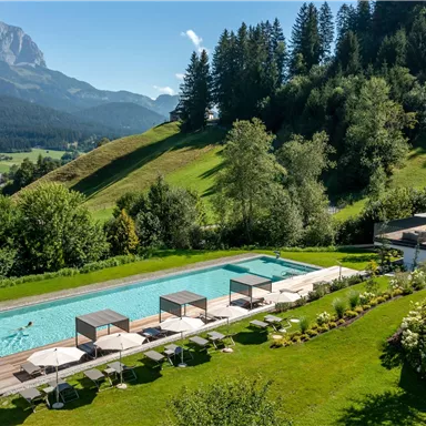 A beautiful outdoor pool surrounded by a green garden and mountains in the background. Relaxing loungers invite you to linger.