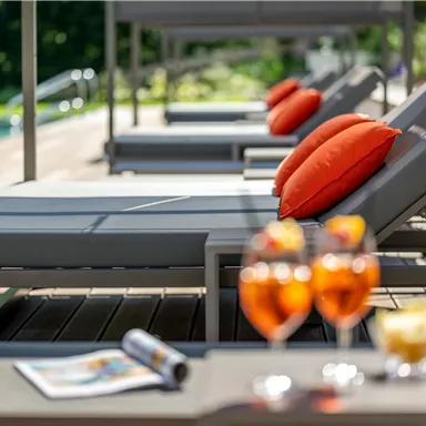 Relaxed atmosphere with loungers and orange cushions by the pool. In the foreground are cocktails and snacks on a table.