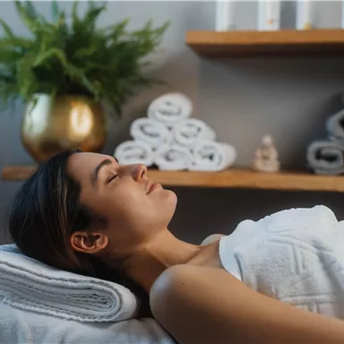 A relaxed woman is lying on a lounge chair, covered with a towel. In the background, folded towels and a houseplant can be seen.
