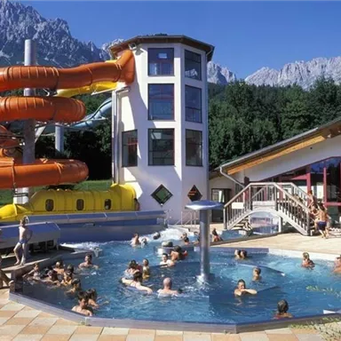 Ein Wasserpark mit einem großen Schwimmbecken und einer Rutsche. Im Hintergrund sind majestätische Berge und Sonnenschein zu sehen.