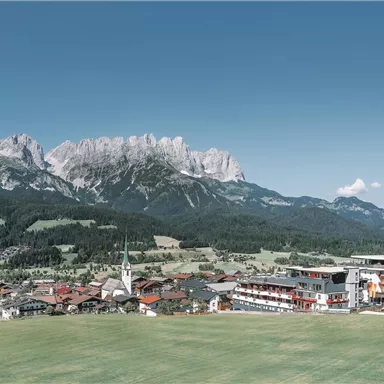 A picturesque mountain landscape with majestic mountains and a small village in the foreground. The sky is clear and blue, and the meadows are green.