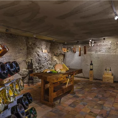 A rustic wine cellar with wooden shelves displaying various wines and other products. In the center stands a large wooden table that invites you to linger.