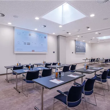 A modern conference room with tables and chairs.  
There is a screen on the wall and plenty of natural light through the skylight.