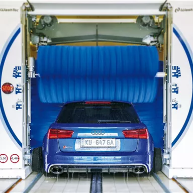 A blue car in a car wash with large blue brushes. The car wash is open and the car is being cleaned.